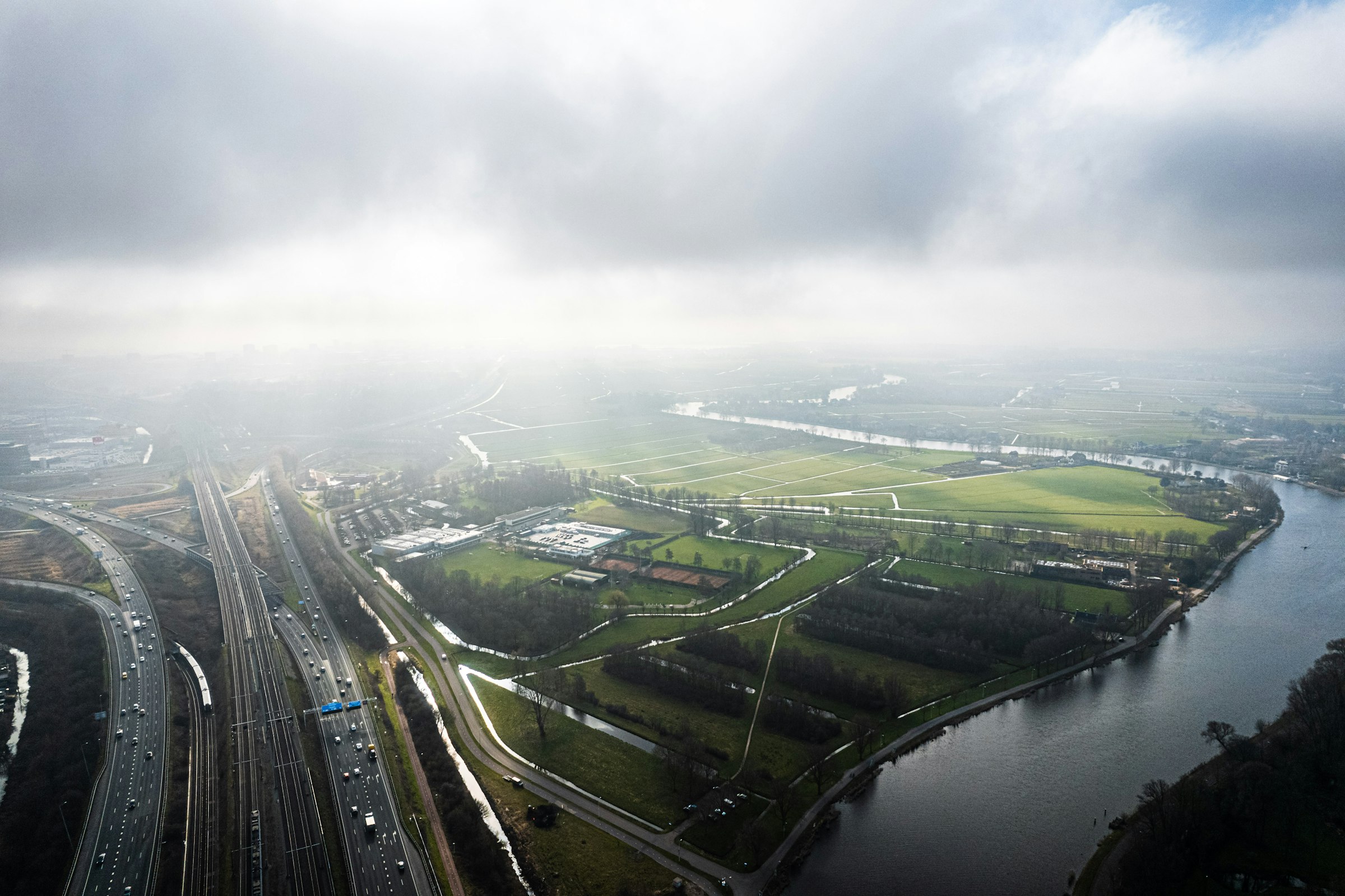 An aerial view of the river Amstel curving past a motorway and green polder land under low mist.