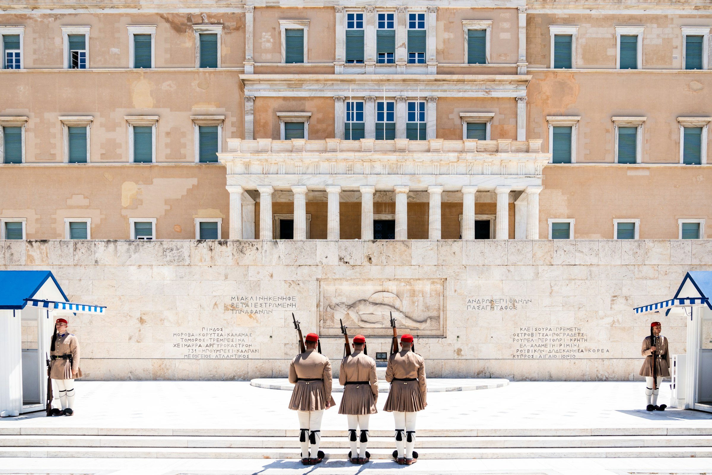 Three Evzones guards in ceremonial uniform marching in front of the Greek Parliament building.