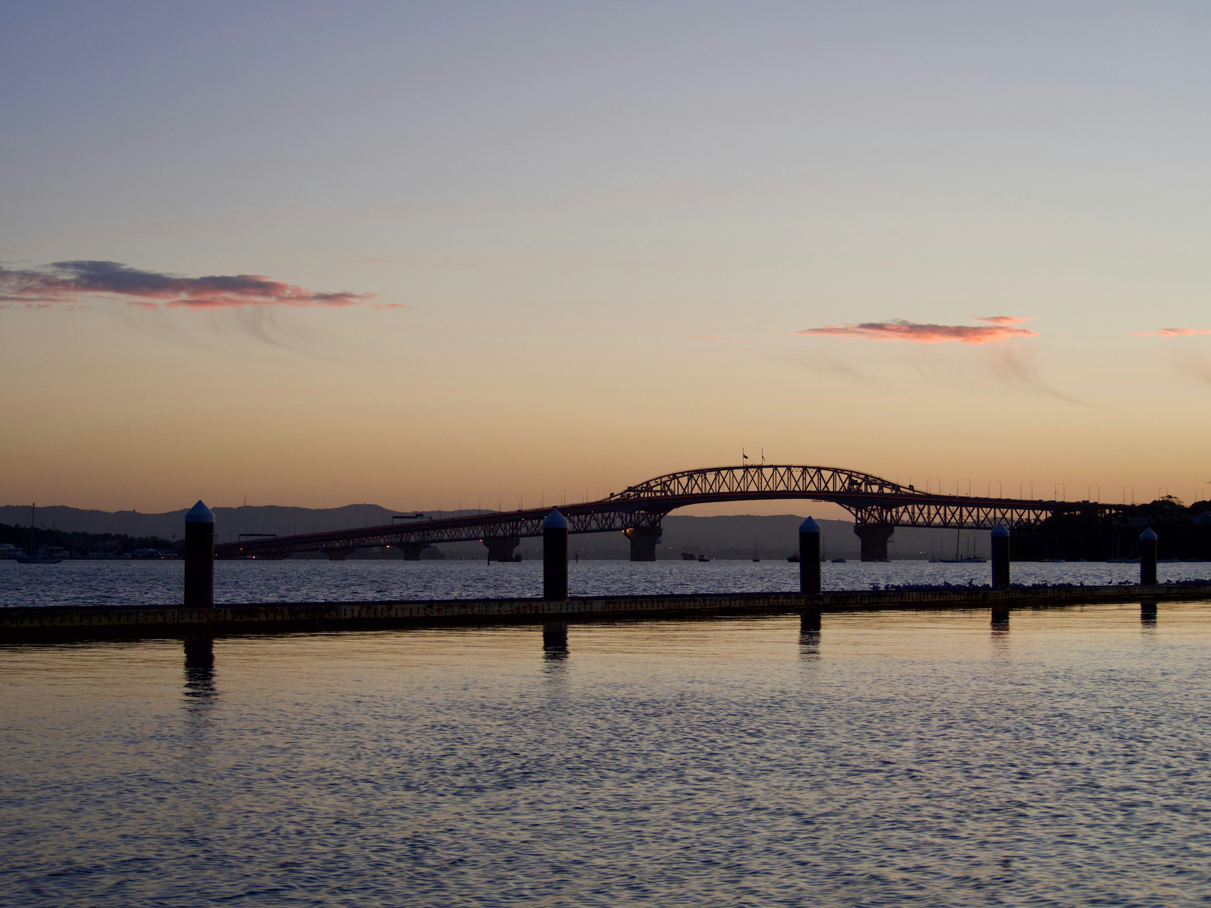 The arch of the Auckland Harbour Bridge in silhouette over still water at sunset, pier posts in the foreground.