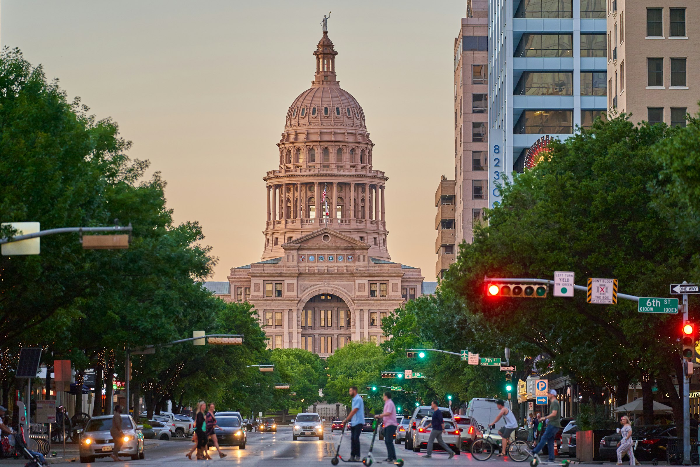 A city avenue with traffic lights and people crossing, leading up to the domed Capitol in warm evening light.