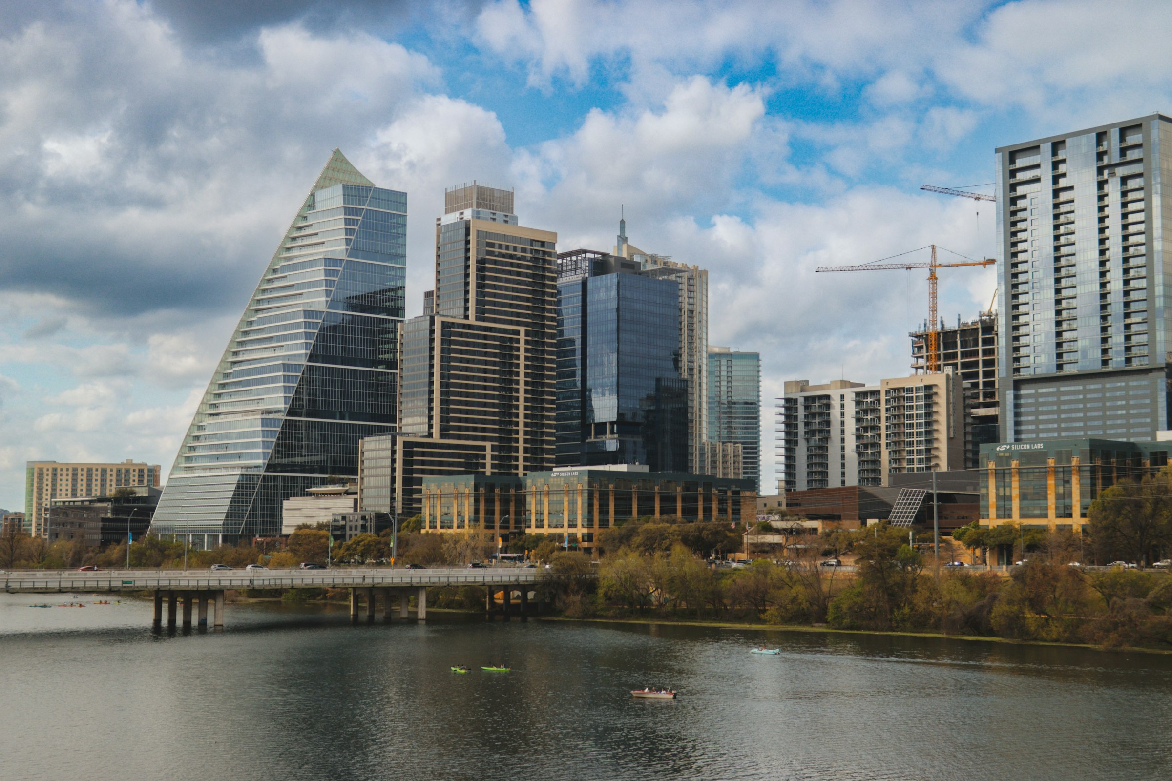 Glass towers, one a tall angled triangle, rising above a calm river lined with trees, under white clouds.