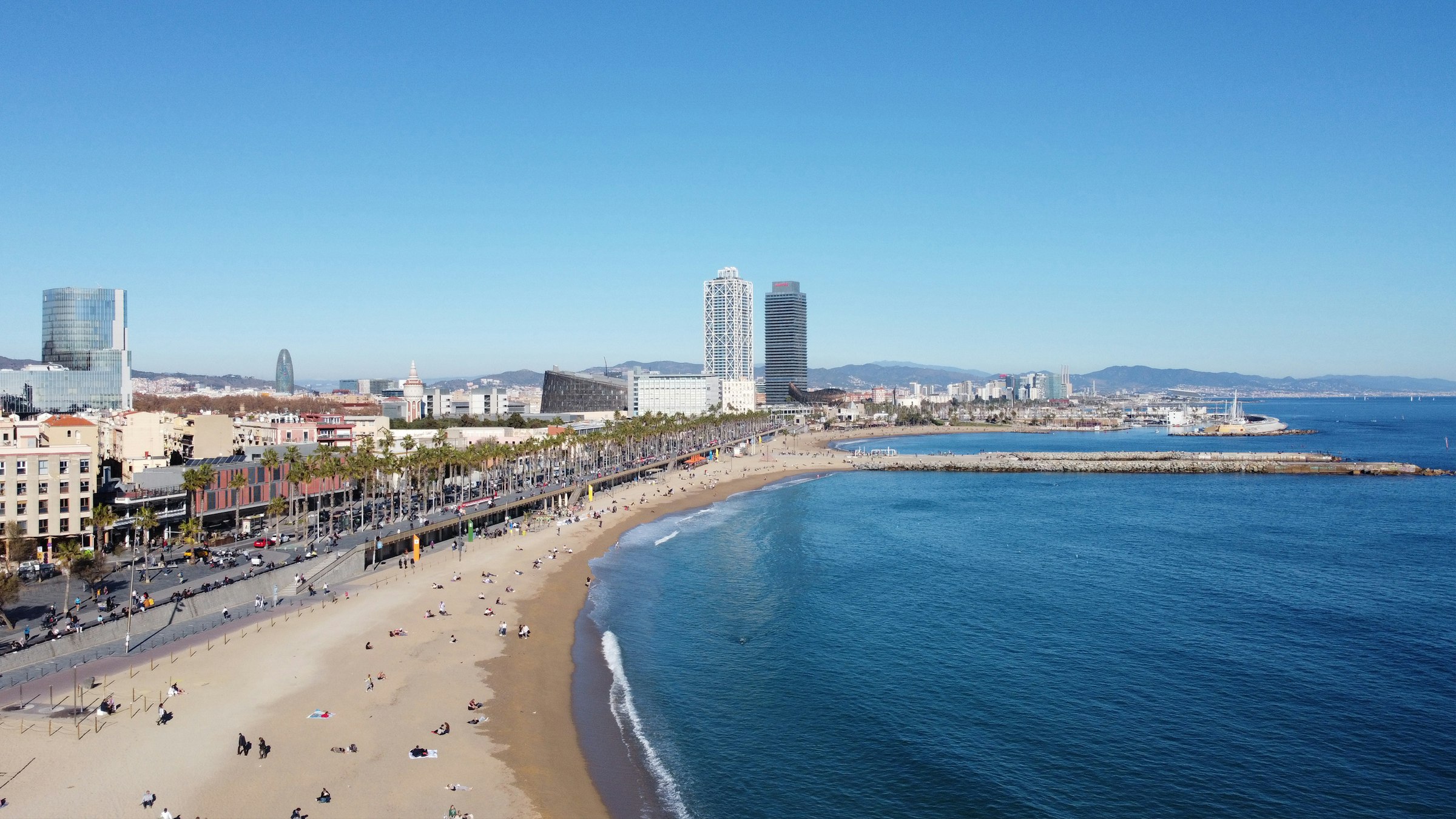 A curving city beach and promenade with two tall towers at the marina beyond.