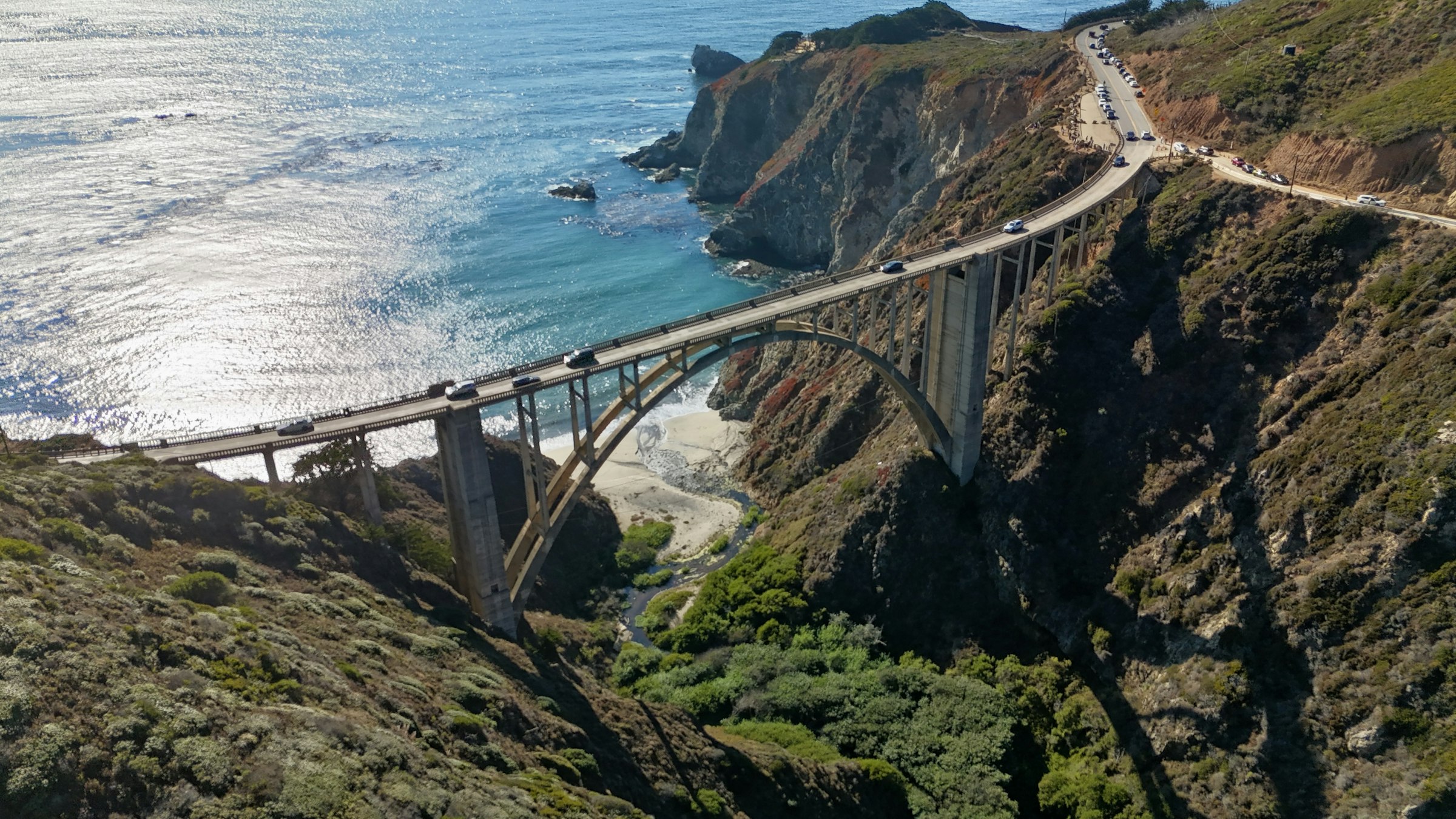 Bixby Creek Bridge from the air, turquoise water in the cove beneath it.