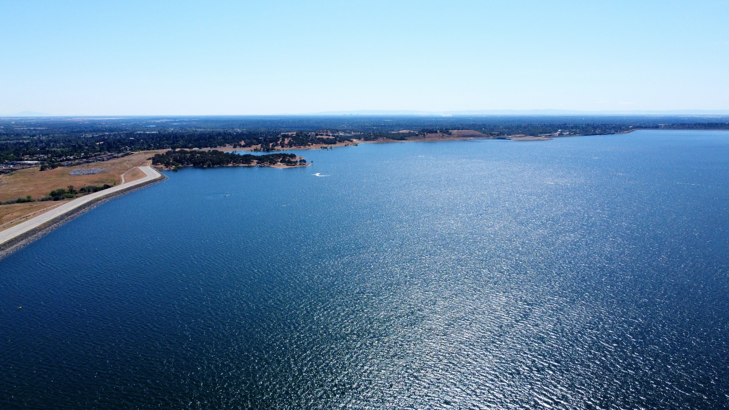 A wide blue lake seen from the air, with a sandy shoreline and wooded hills along the far side.
