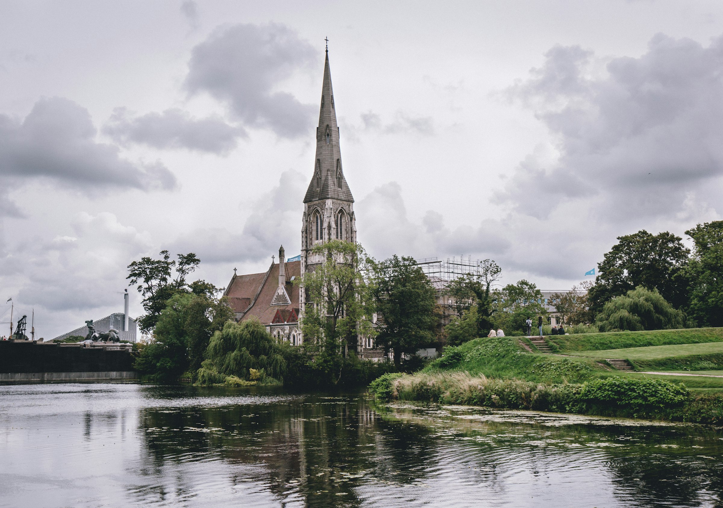 A Gothic stone church with a tall spire beside a pond, under a grey sky.