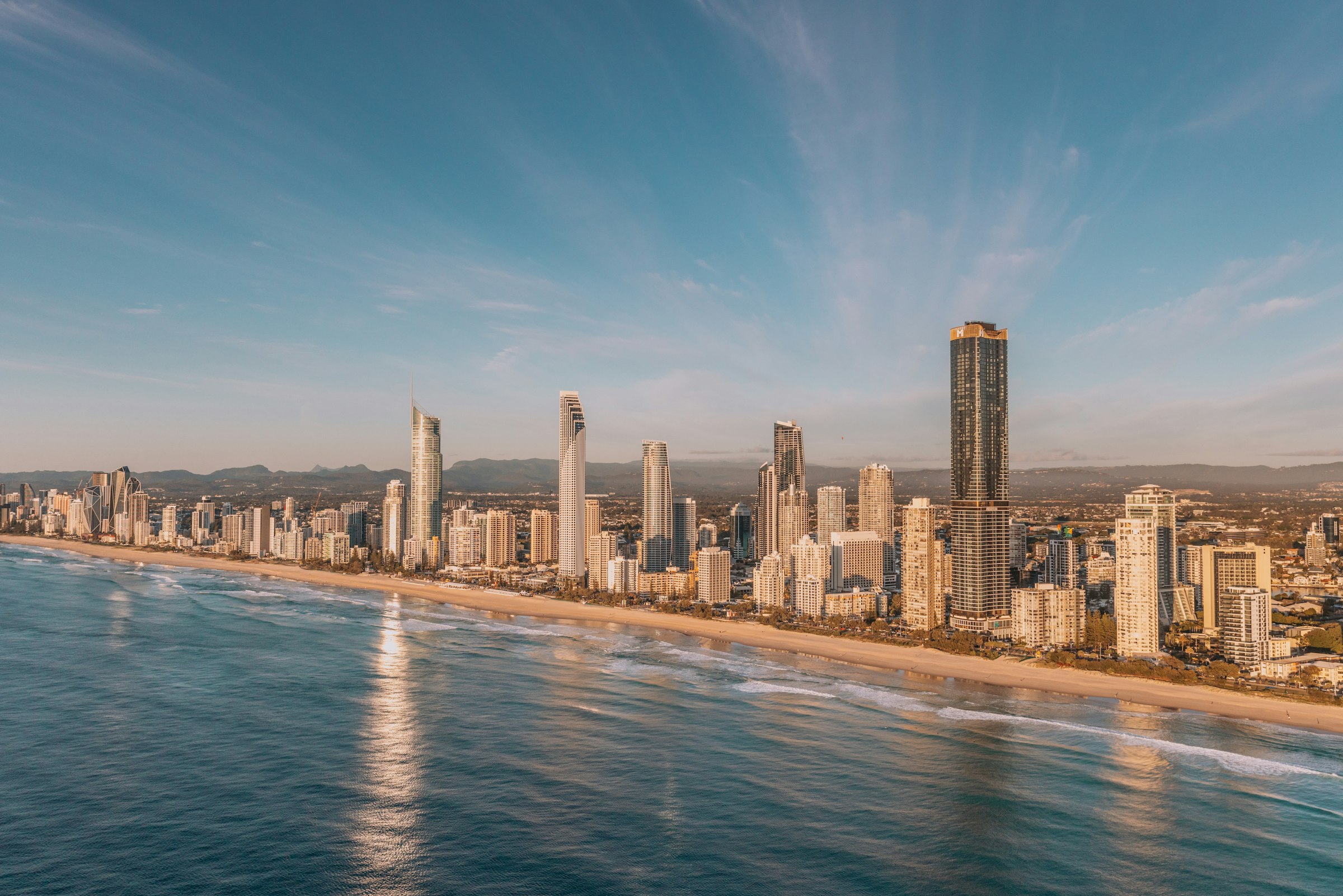 The towers of Surfers Paradise lit by low sun above a long beach and calm sea.