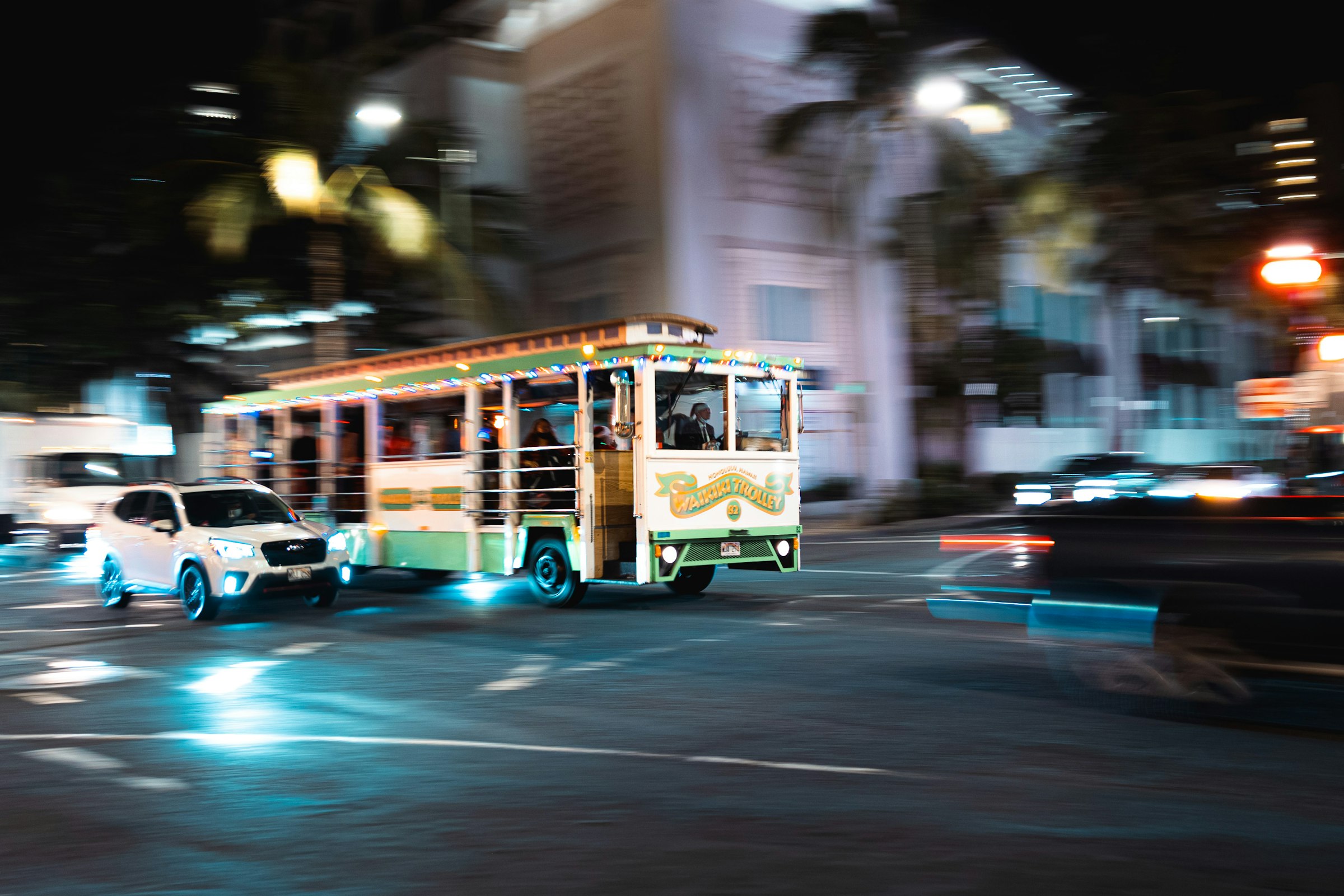 A Waikiki trolley bus moving along a lit city street at night, blurred by a long exposure.