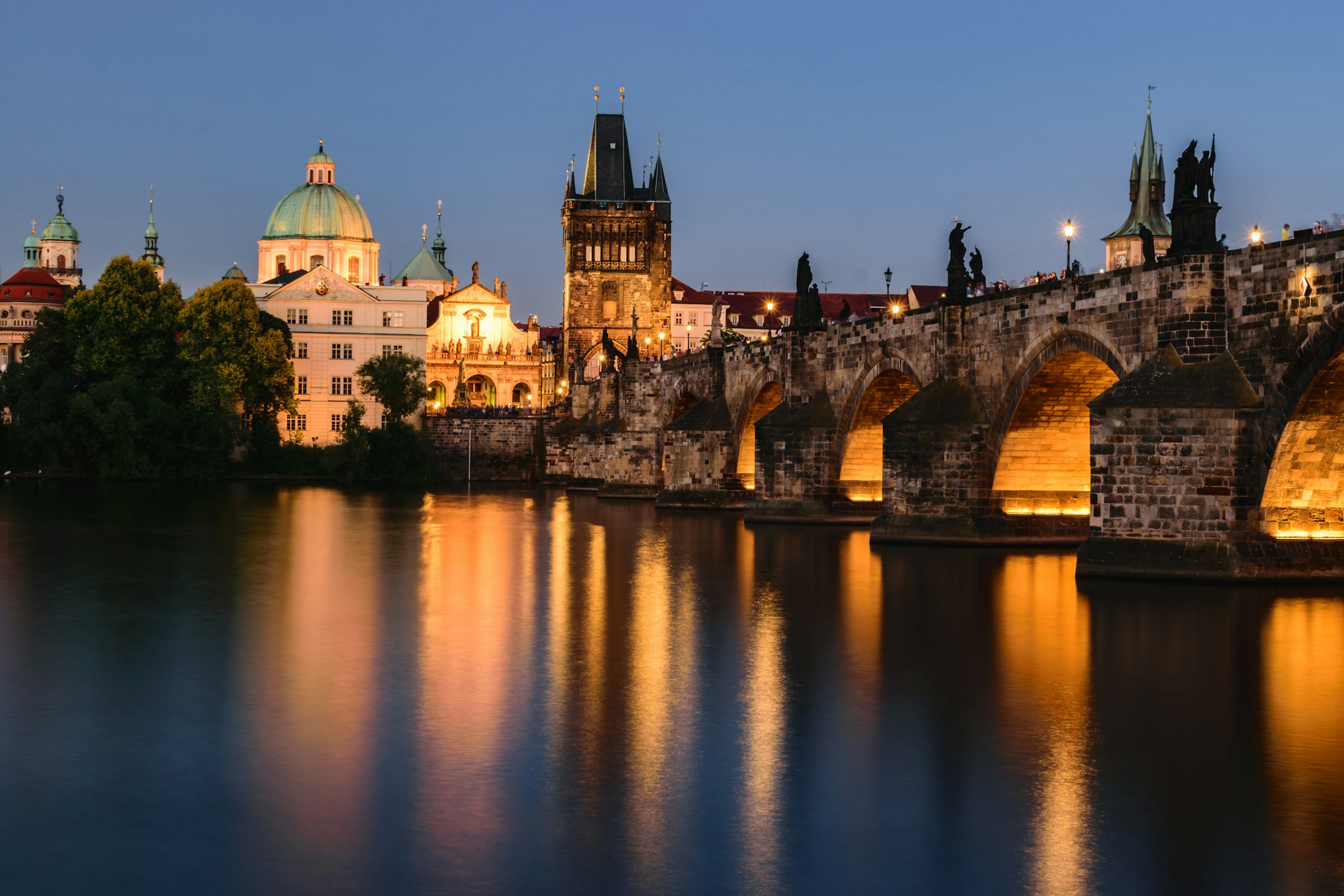 The stone arches of the Charles Bridge lit up at dusk and reflected in the river, with a Gothic tower at the end.