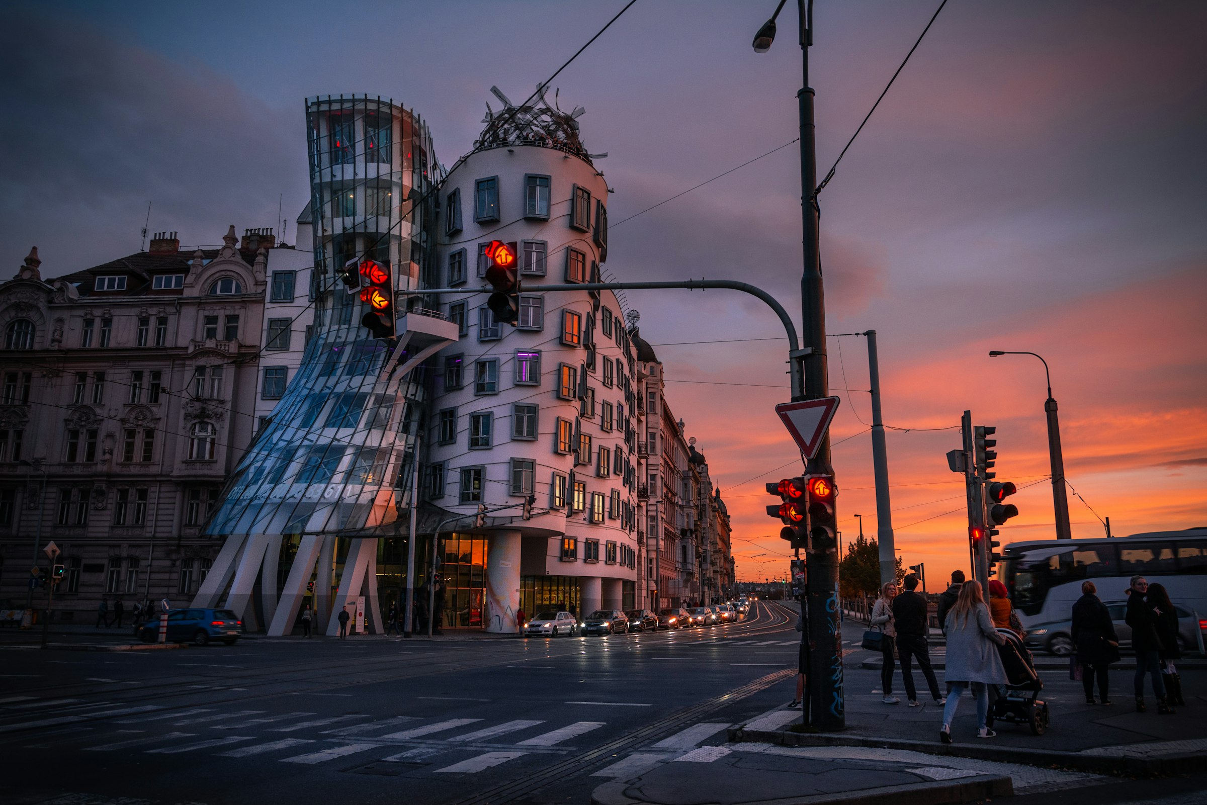 The curved glass and concrete Dancing House on a street corner at sunset, with a tram signal in front.