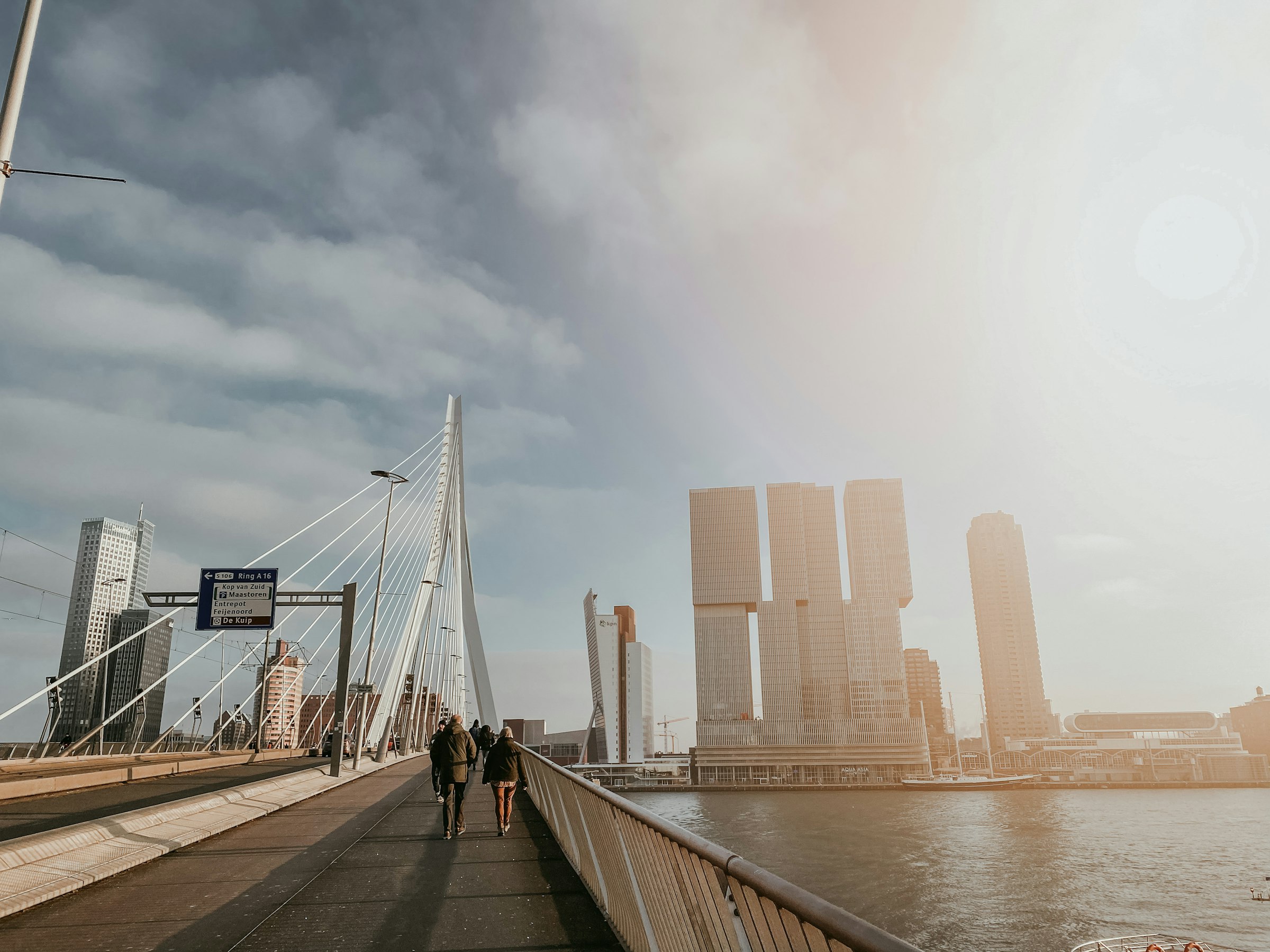 People walking across a bridge deck under tall white cables, with high-rise towers across the river in hazy light.