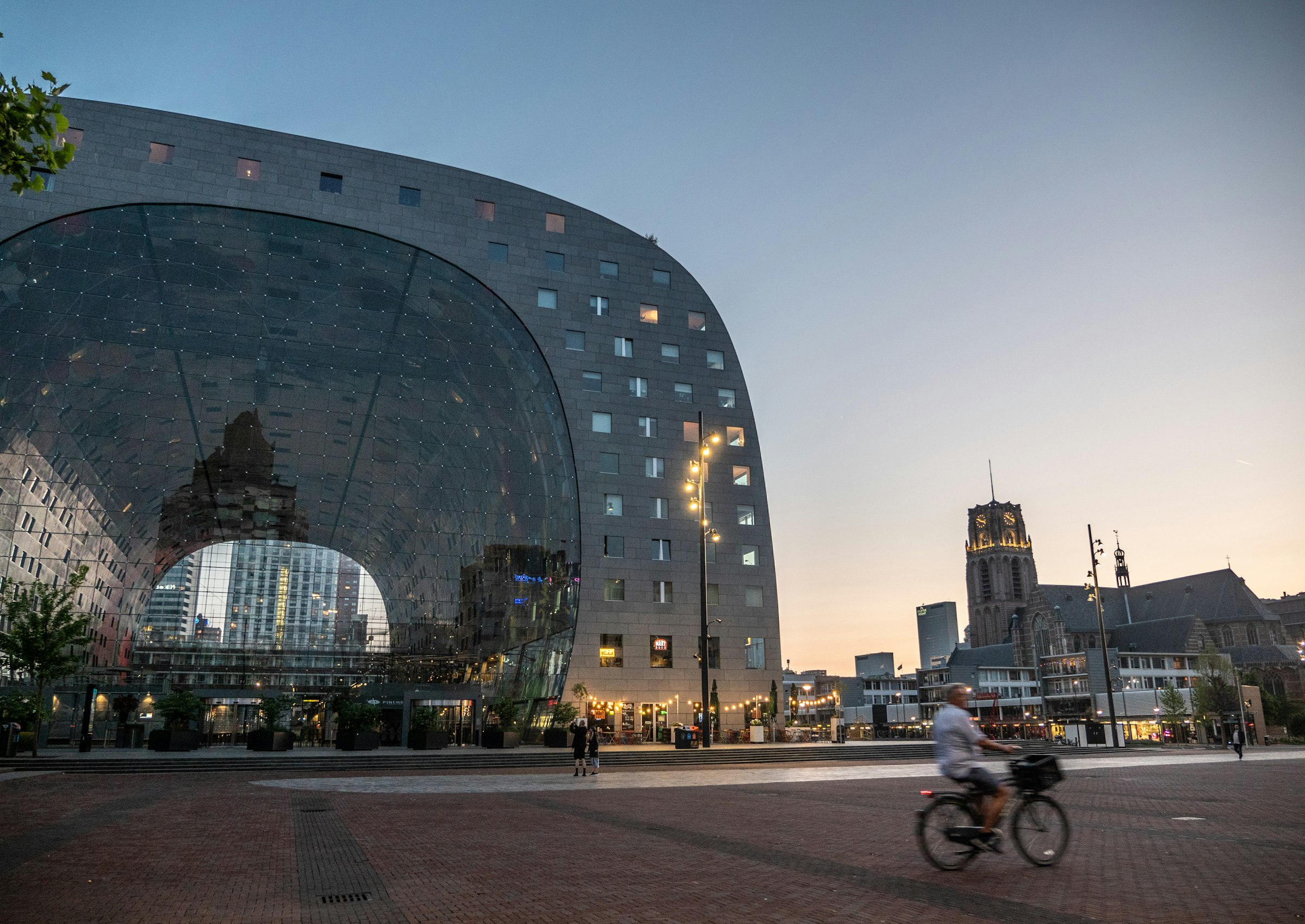 The great arched market hall of Rotterdam, its glass end wall lit from inside, with a cyclist crossing the square.