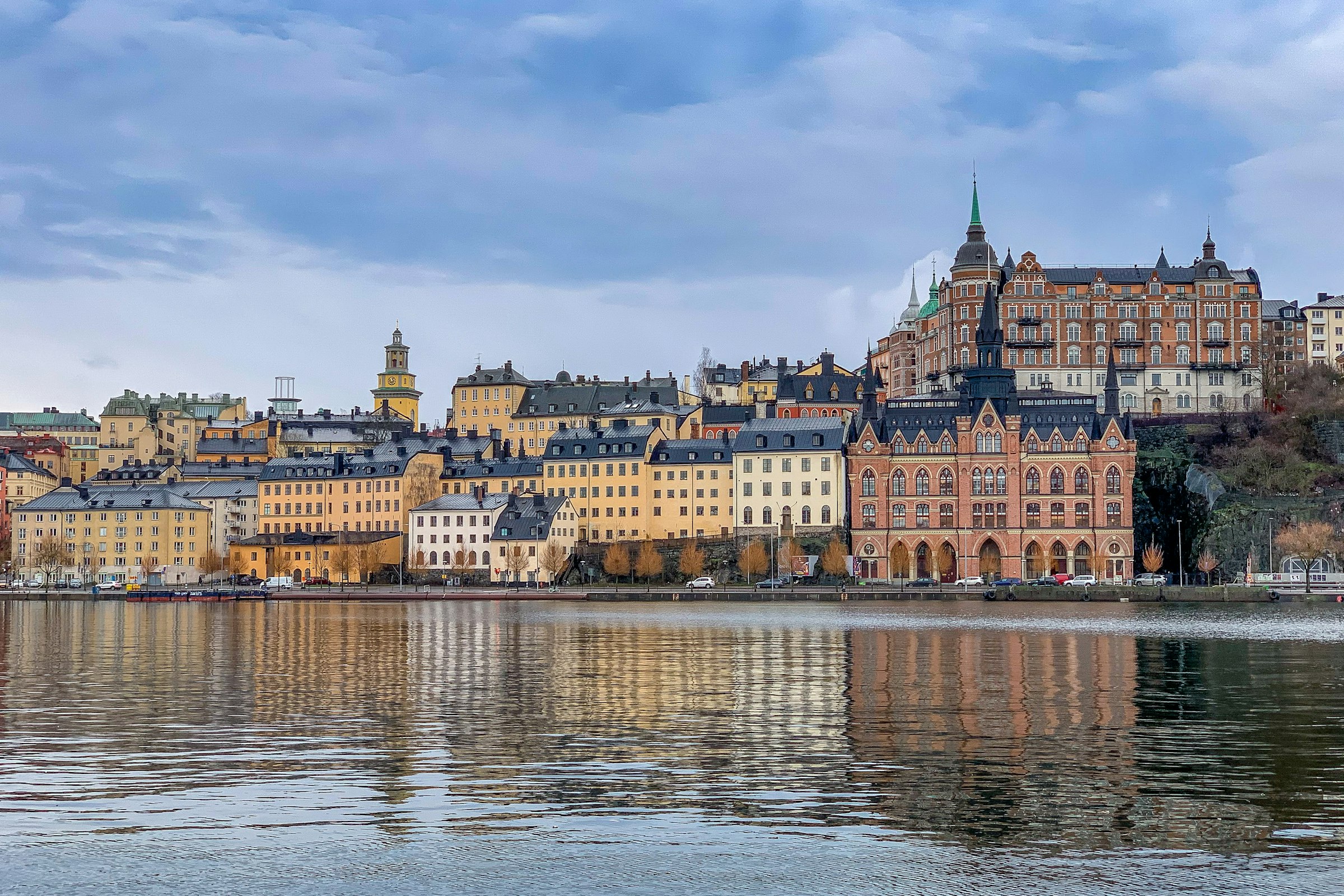 Tall ochre and red buildings along a waterfront, reflected in calm water under a pale sky.