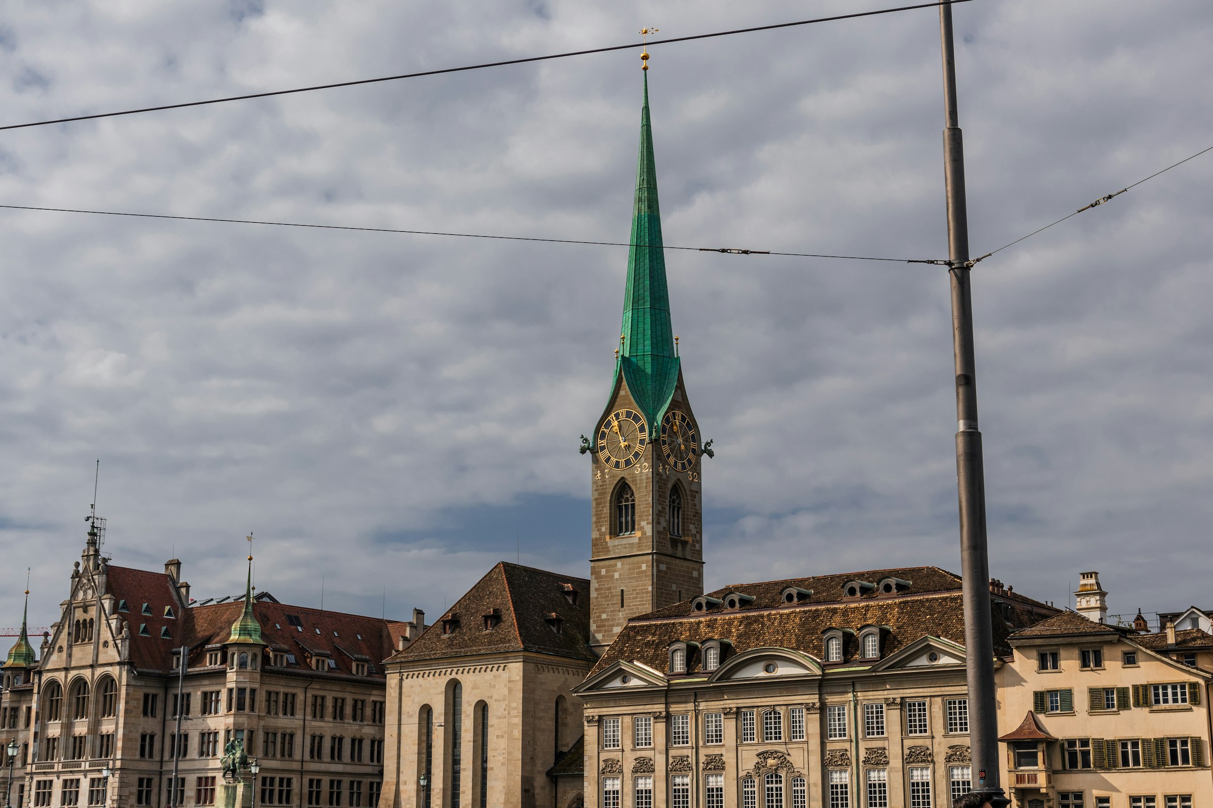 The Fraumünster church with its tall green spire, beside guild houses, under a grey sky.