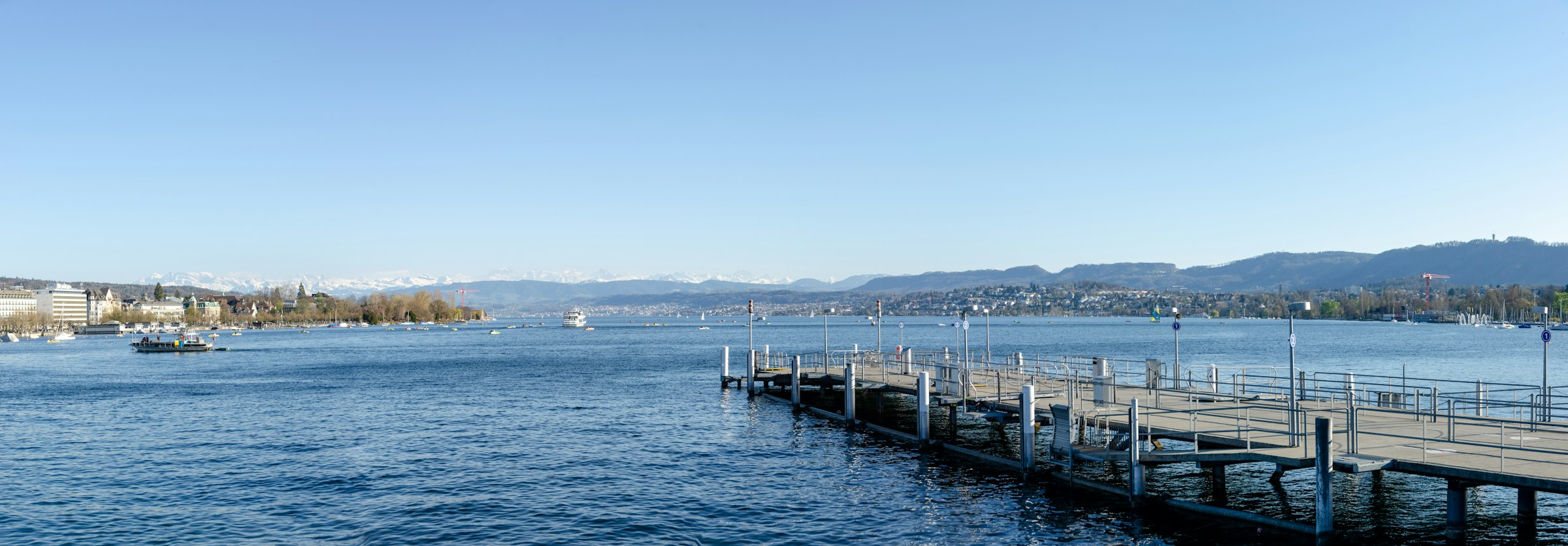 A long wooden pier stretching into a calm blue lake, with low hills and distant mountains beyond.