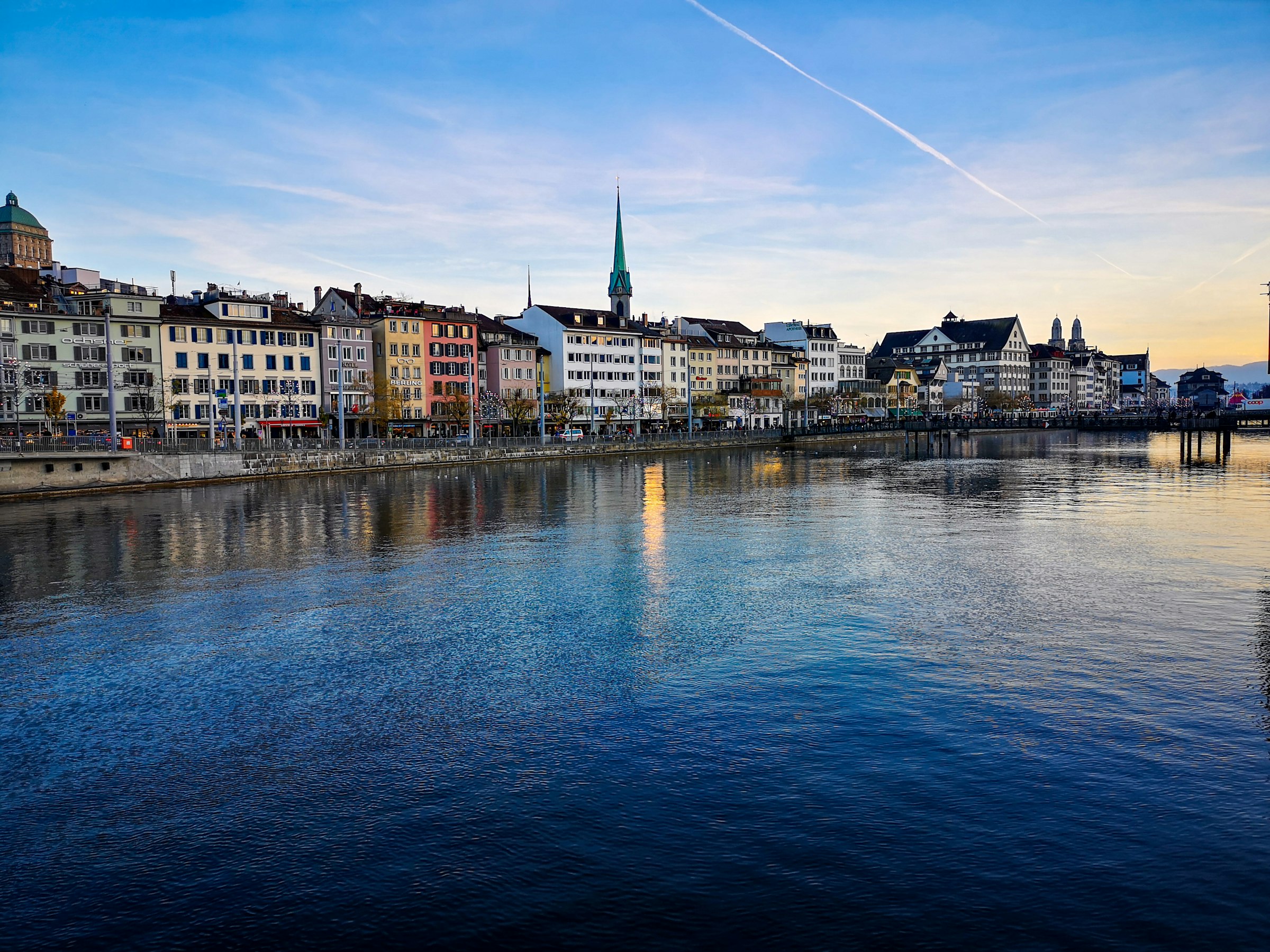 Old town houses and a church spire along a river in soft morning light.